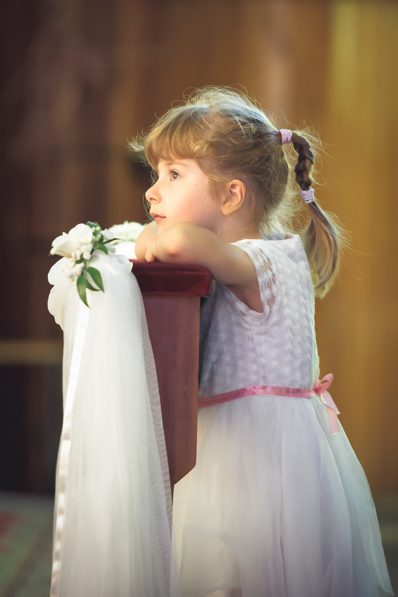 Dziewczynka w białej sukience z różową wstążką, oparta o ozdobiony kwiatami i tiulem klęcznik, zamyślona patrzy w dal, w tle rozmyte wnętrze kościoła.