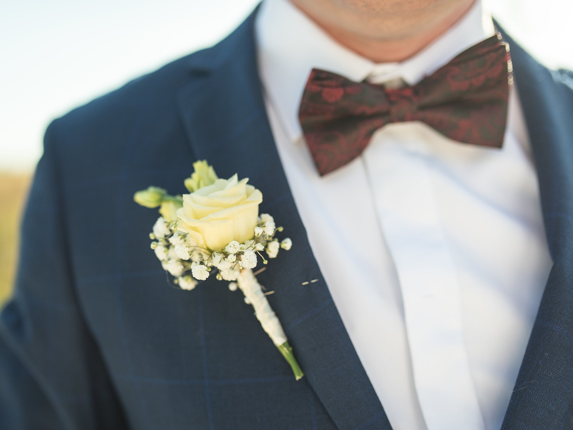 Detal portretu pana młodego w granatowym garniturze z bordową muchą i butonierką z kremowej róży i gipsówki.