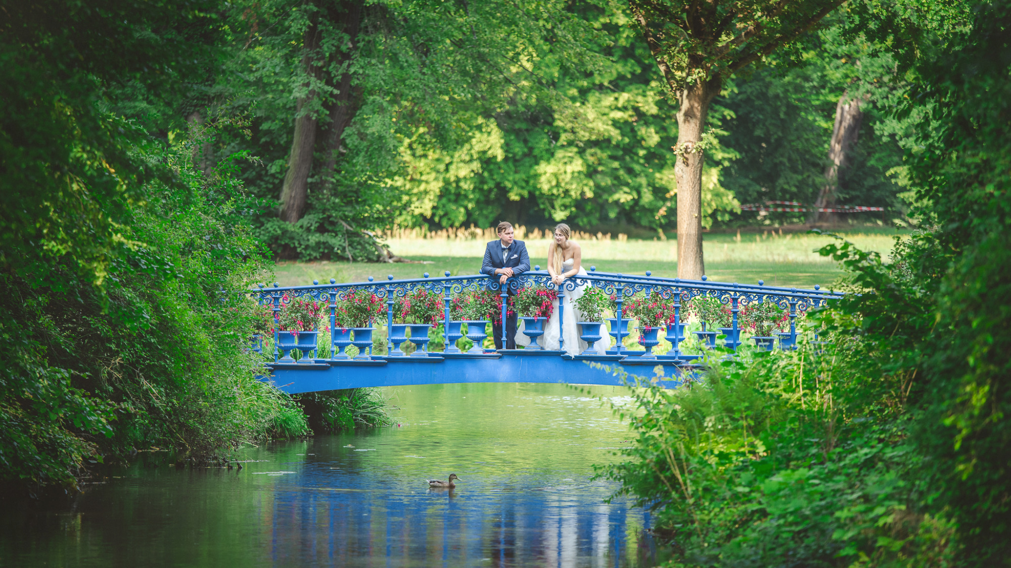 Para młoda w strojach ślubnych stoi na niebieskim moście ozdobionym kwiatami, z kaczką pływającą w wodzie pod mostem.