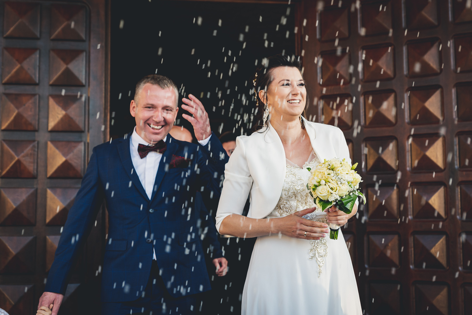 Uśmiechnięta para młoda wychodzi z kościoła obsypywana ryżem, panna młoda w białej sukni i bolerku trzyma bukiet, pan młody w granatowym garniturze z bordową muszką.