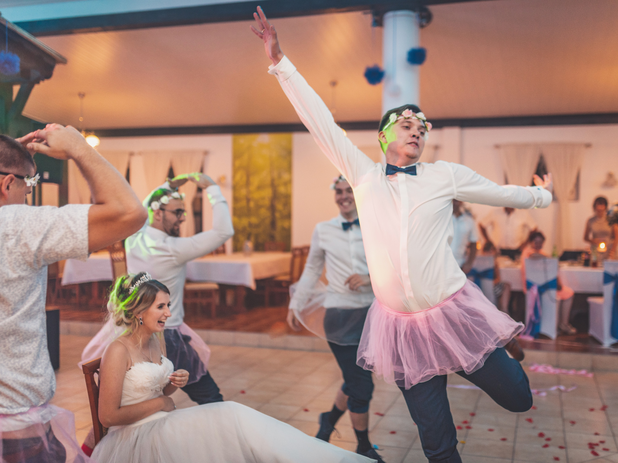Panna młoda siedzi na krześle podczas zabawy oczepinowej z mężczyznami w tiulowych spódniczkach i wiankach na głowach.