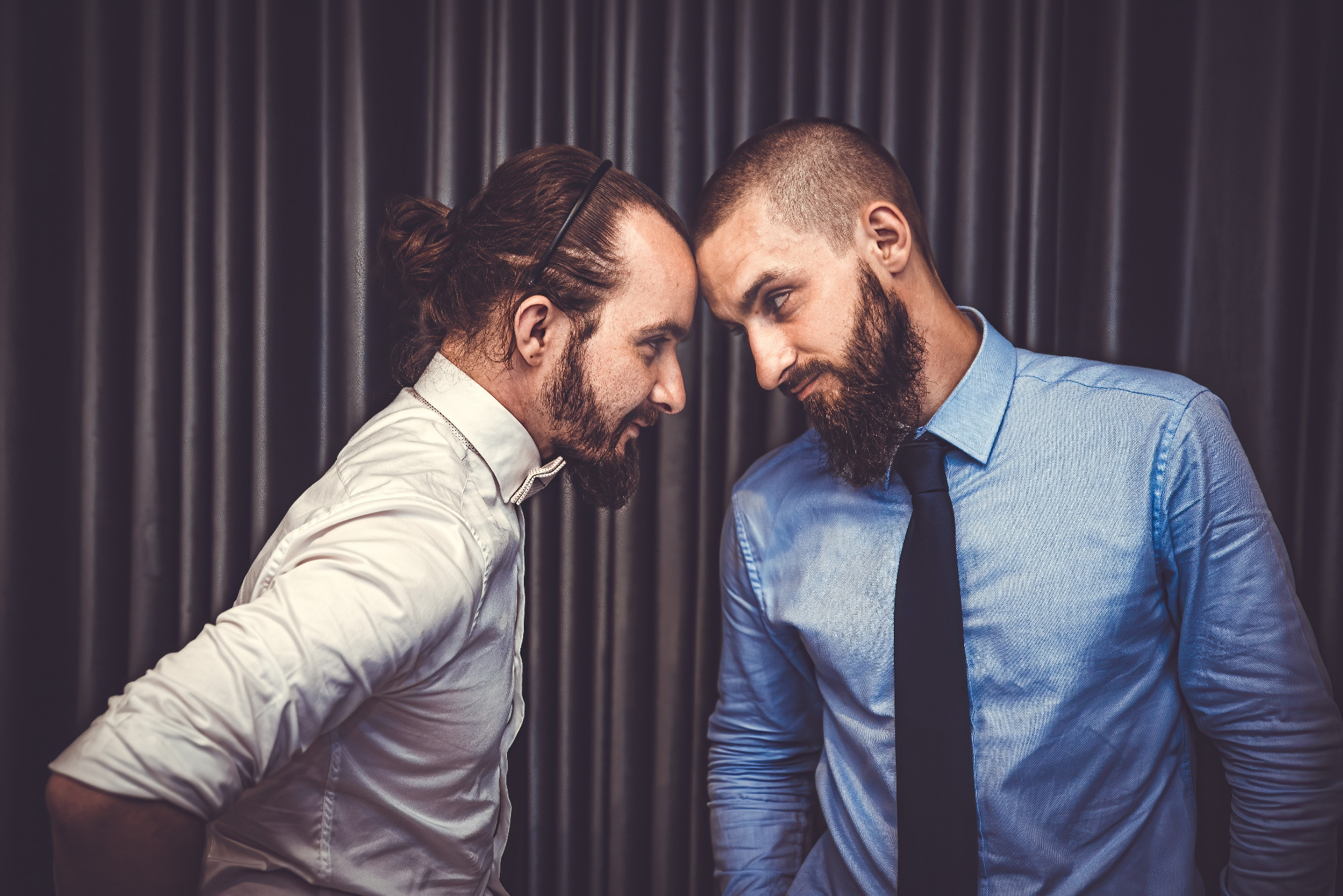 Portret dwóch brodatych mężczyzn w koszulach i krawatach, stojących twarzami do siebie na tle ciemnej zasłony.