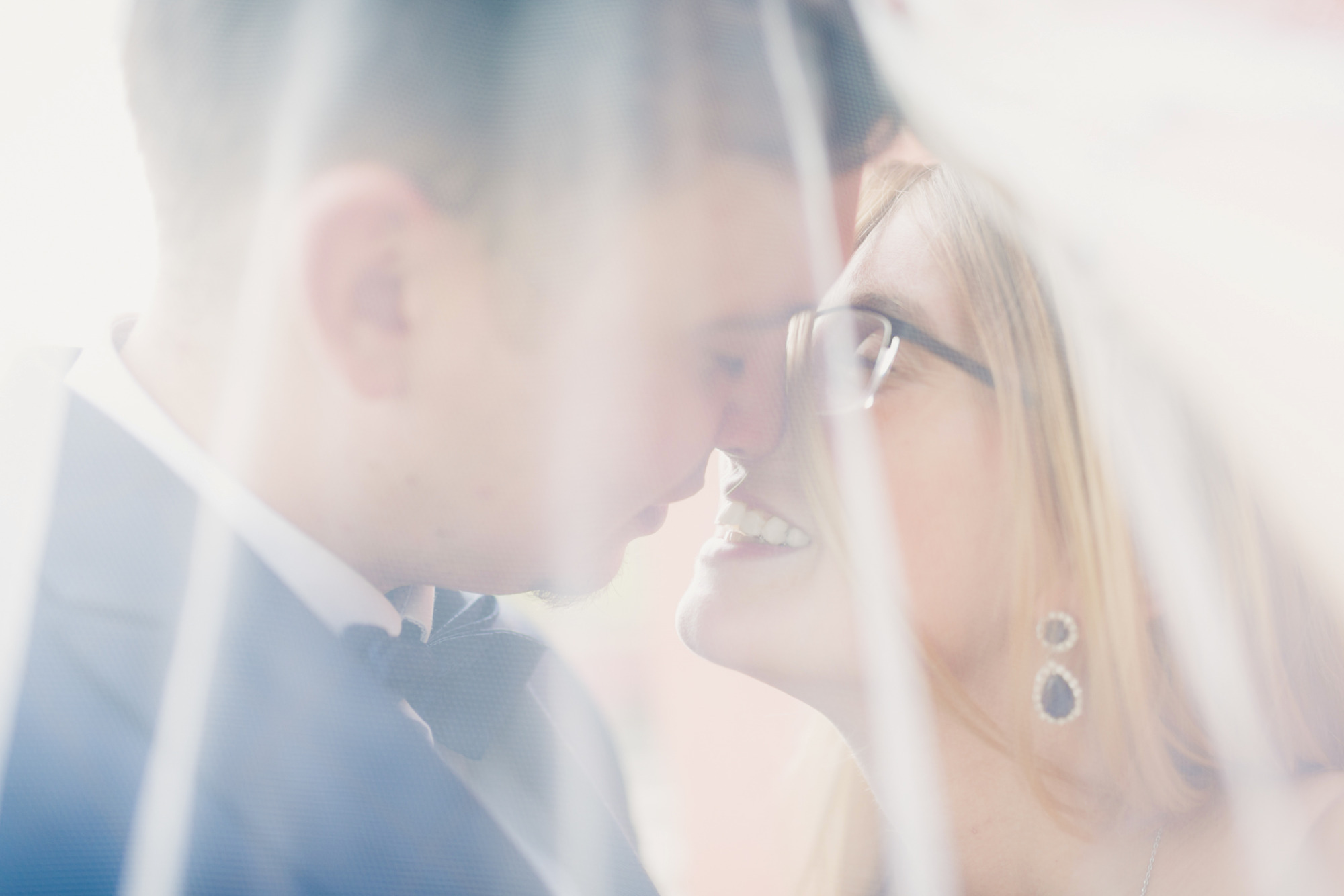 Para młoda w zbliżeniu, panna młoda w okularach, widoczna przez delikatny welon, z uśmiechem spogląda na pana młodego.