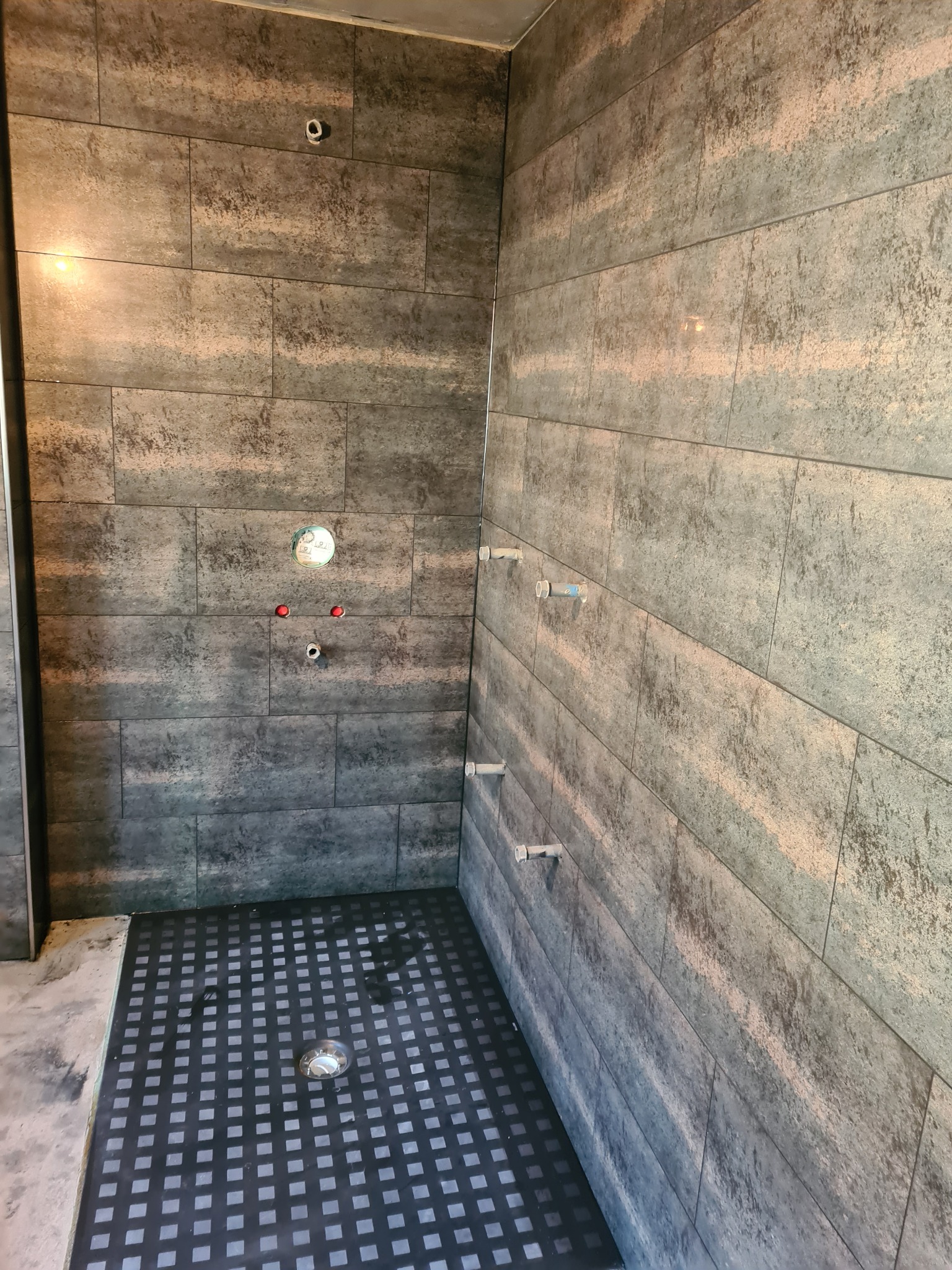 Wnętrze kabiny prysznicowej w trakcie wykańczania, z ciemnymi, strukturalnymi płytkami na ścianach i podłogą z kratką odpływową. Widoczne wystające rury i otwory na baterię.