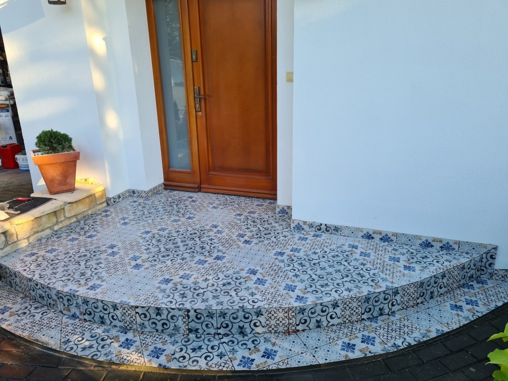 Wyłożone wzorzystymi płytkami schody wejściowe do domu z drewnianymi drzwiami, z doniczką z rośliną po lewej stronie.