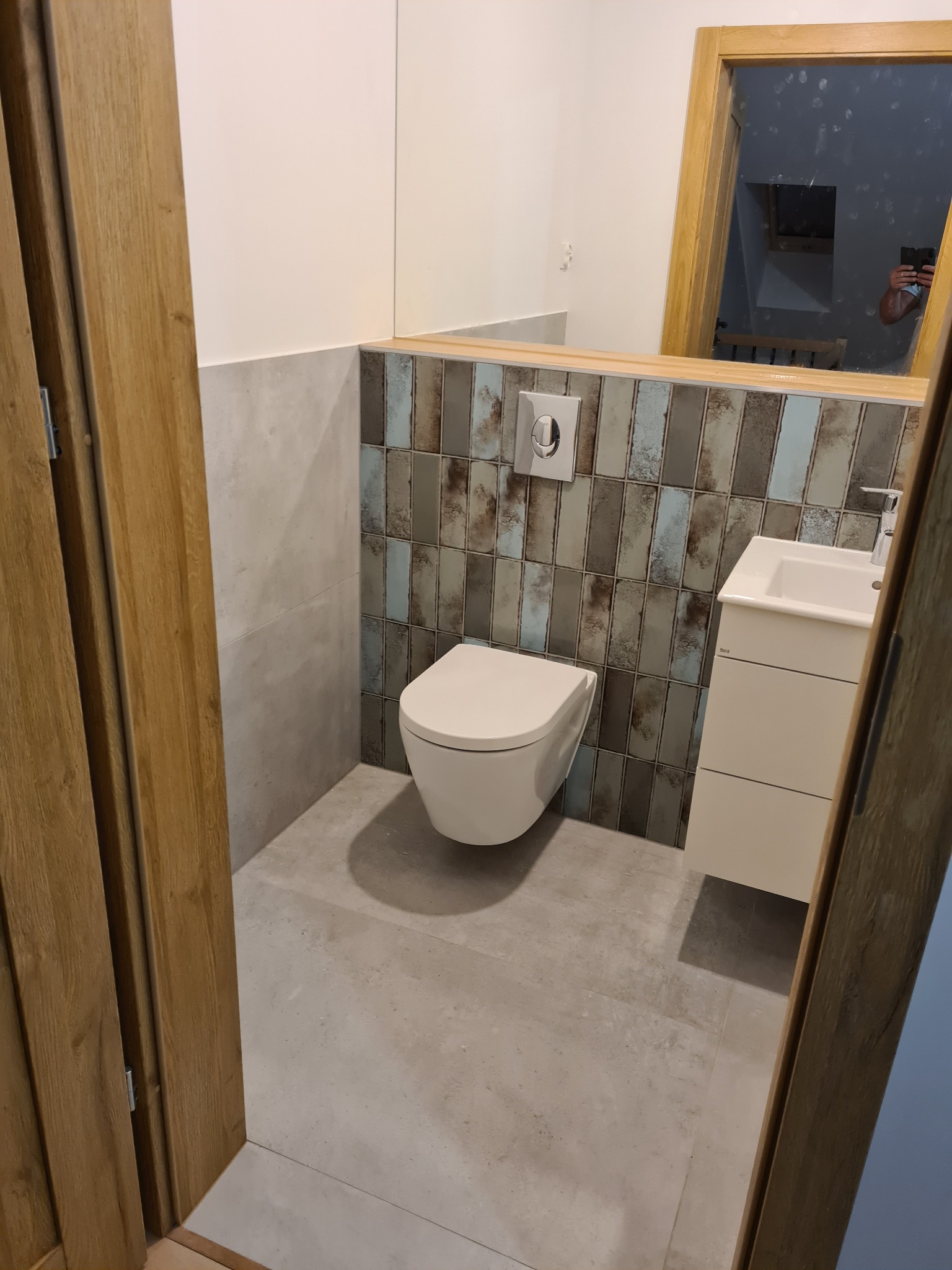 Nowoczesna toaleta podwieszana w łazience z płytkami w odcieniach szarości i błękitu, widziana zza drewnianej futryny drzwiowej.