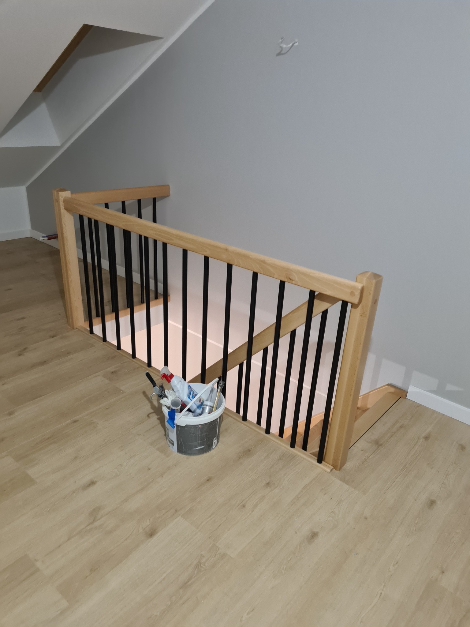 Poddasze po odnowieniu: jasne panele podłogowe, świeżo pomalowane szare ściany, drewniana balustrada schodów z czarnymi elementami, wiadro z farbami i narzędziami malarskimi w rogu.