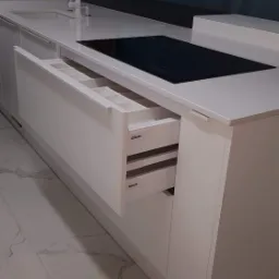 Biała kuchnia z otwartymi szufladami marki Blum, widoczna płyta indukcyjna i bateria umywalkowa, marmurowa podłoga.