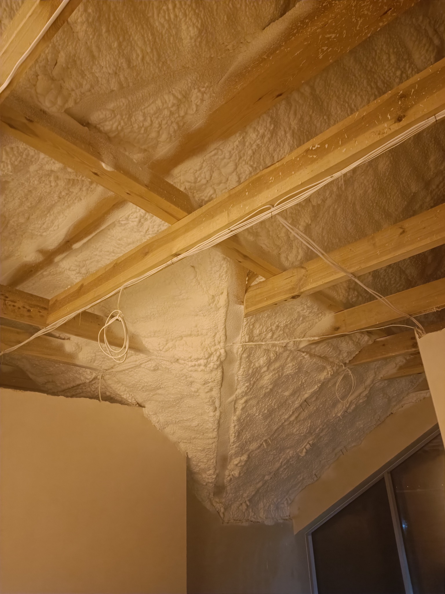 Poddasze ocieplone pianką, widoczne drewniane krokwie i instalacja elektryczna, szorstka faktura izolacji.