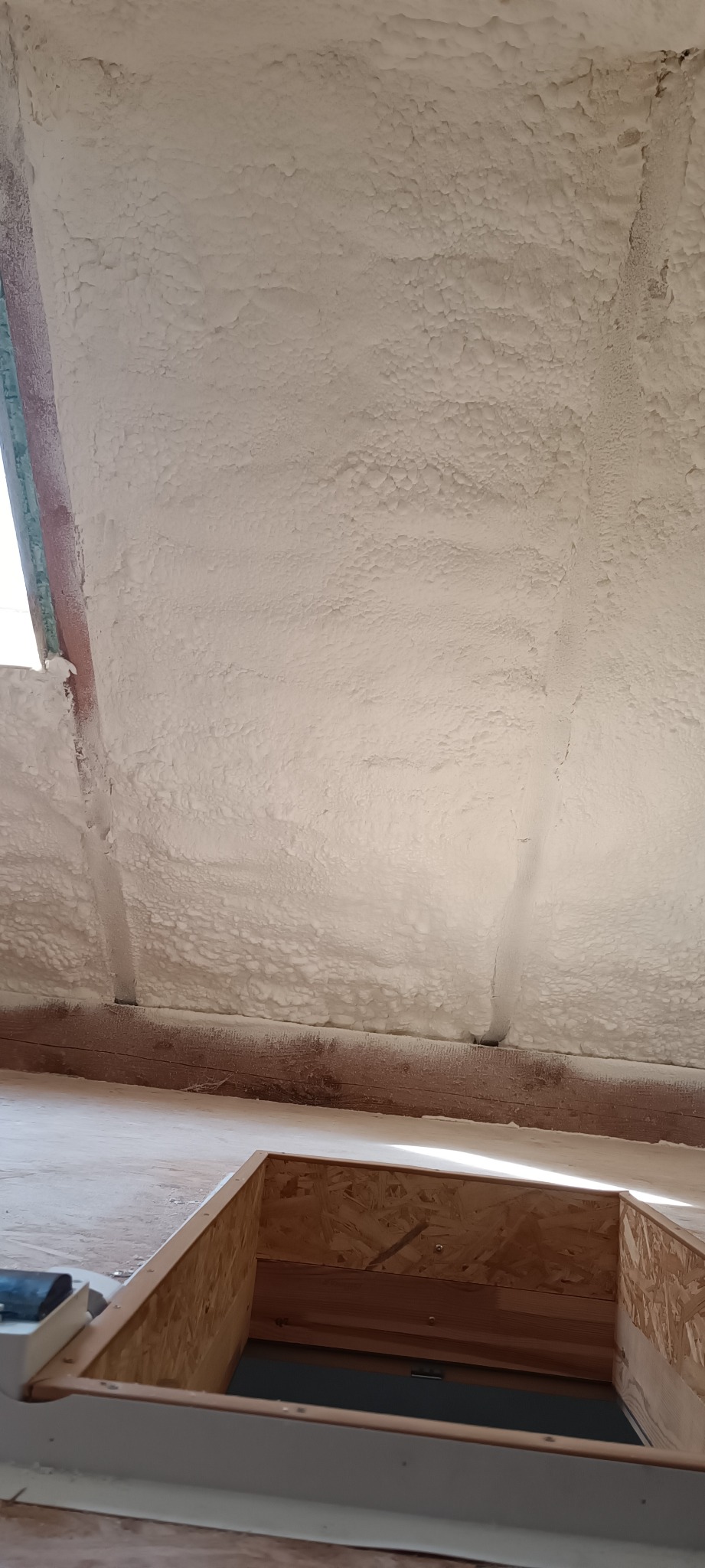 Poddasze po natrysku pianki izolacyjnej, widoczna drewniana konstrukcja i okno dachowe w trakcie montażu.