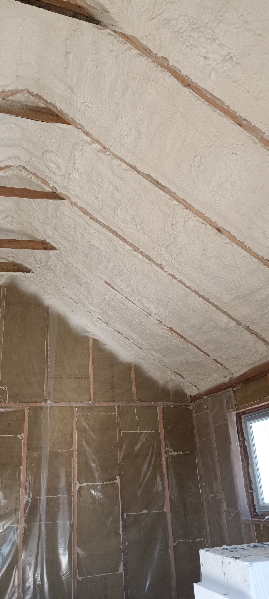 Poddasze z drewnianą konstrukcją pokryte grubą warstwą jasnej pianki izolacyjnej, ściany wyłożone wełną mineralną pod przezroczystą folią, widoczne fragmenty okna.