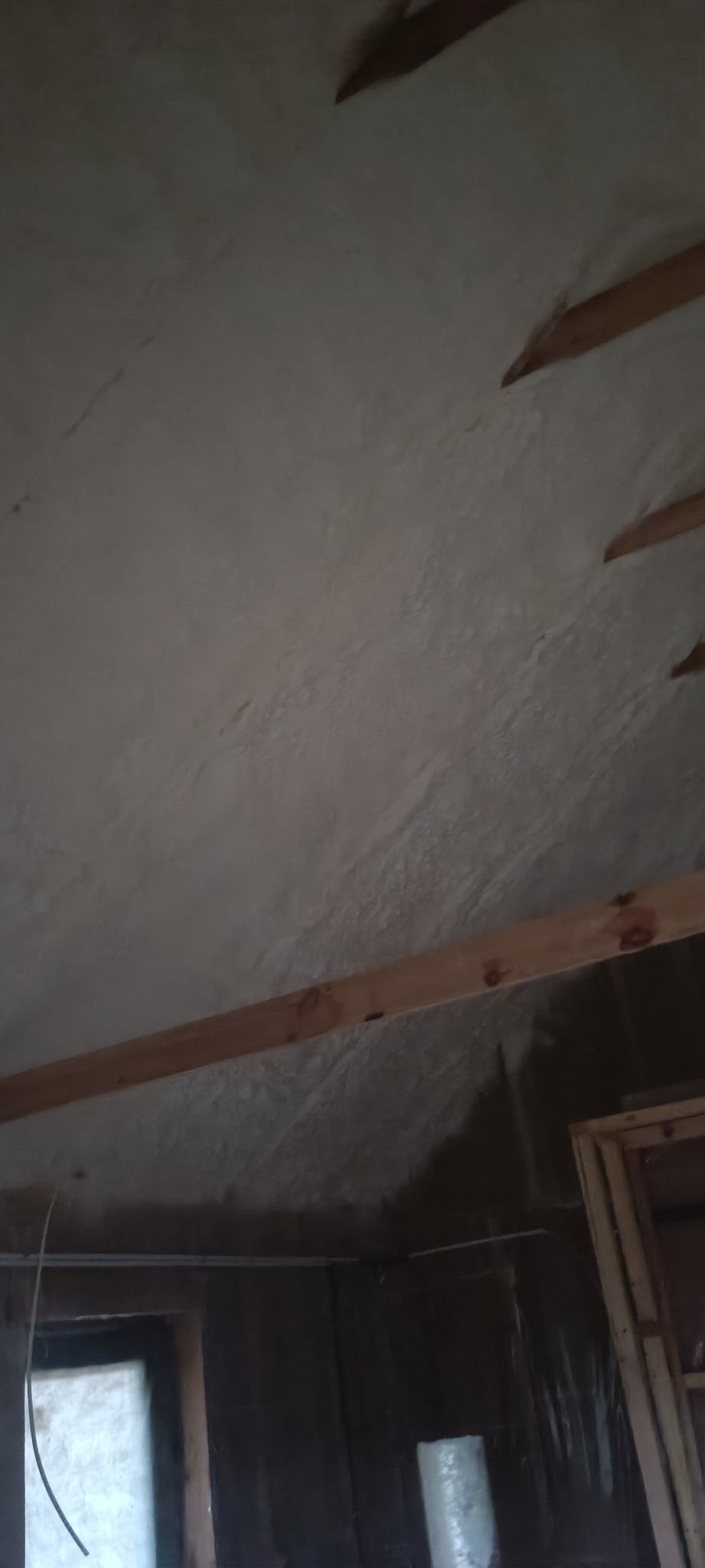 Poddasze z widoczną drewnianą konstrukcją i nierównomiernie nałożoną szarą pianą izolacyjną, widoczne okno w tle.