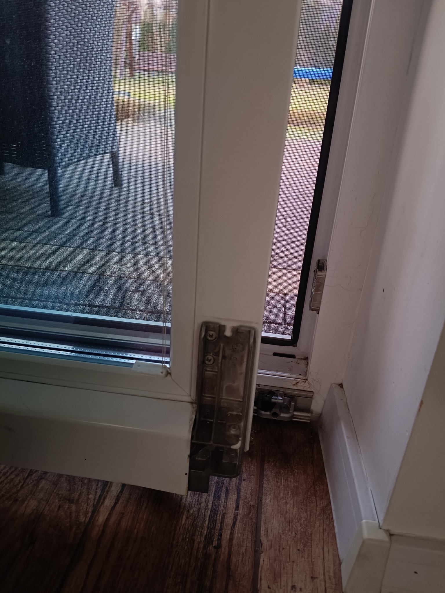 Otwarty fragment białej ramy okiennej z widocznym zawiasem i siatką moskitiery. Widok na zewnątrz przez okno. Fragment podłogi w dolnej części kadru.
