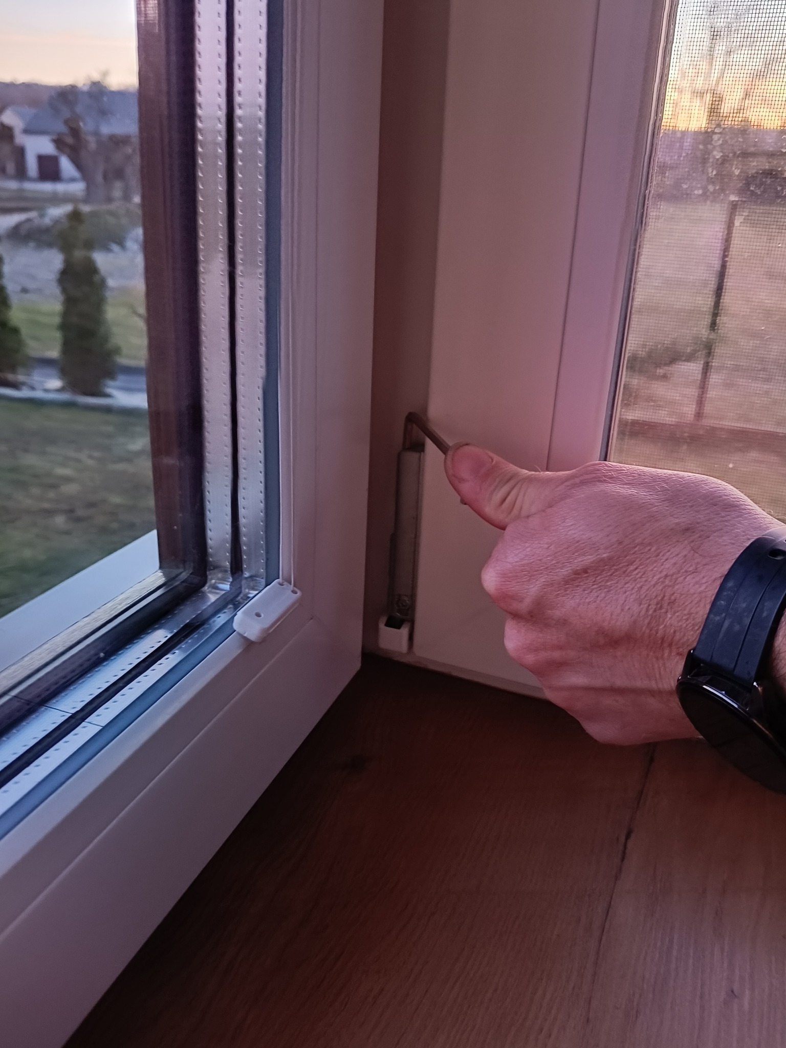 Regulacja okna PCV przy użyciu klucza imbusowego. Widoczna moskitiera w oknie i fragment dłoni z zegarkiem. Ujęcie z bliska, z perspektywy osoby wykonującej regulację.