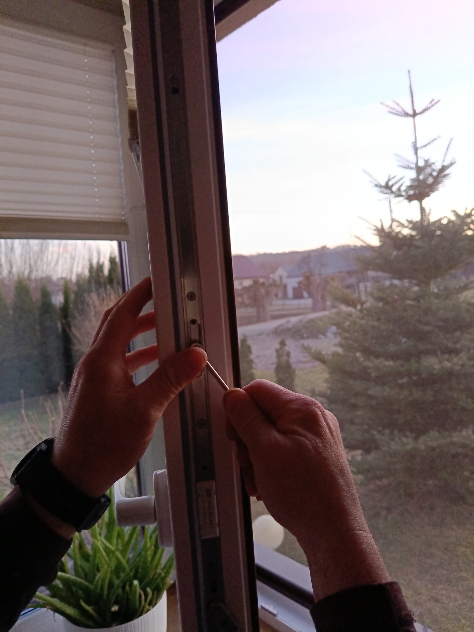 Regulacja okna PCV przy użyciu śrubokręta, widok z bliska na mechanizm okienny, zieleń za oknem i roleta w tle. Dłonie w trakcie pracy.