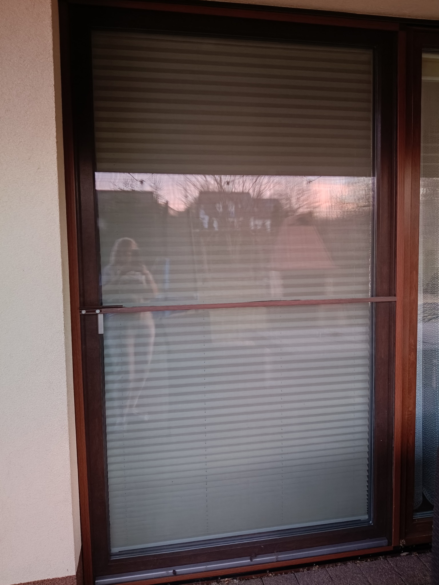 Okno z plisowaną moskitierą w brązowej ramie, widok z zewnątrz. Delikatne odbicie otoczenia na powierzchni siatki. Widoczny mechanizm blokujący moskitierę.