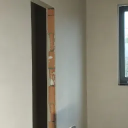 Wnętrze w stanie surowym po tynkowaniu maszynowym, widoczny fragment ściany z ościeżnicą drzwiową w trakcie obróbki, surowy betonowy sufit i lampa budowlana na trójnogu.