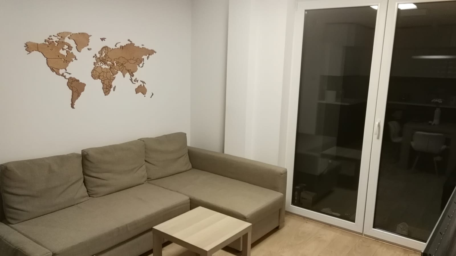 Jasny salon z beżową sofą narożną, niskim stolikiem kawowym i drewnianą mapą świata na białej ścianie, widok na zaciemnioną jadalnię przez przeszklone drzwi balkonowe.