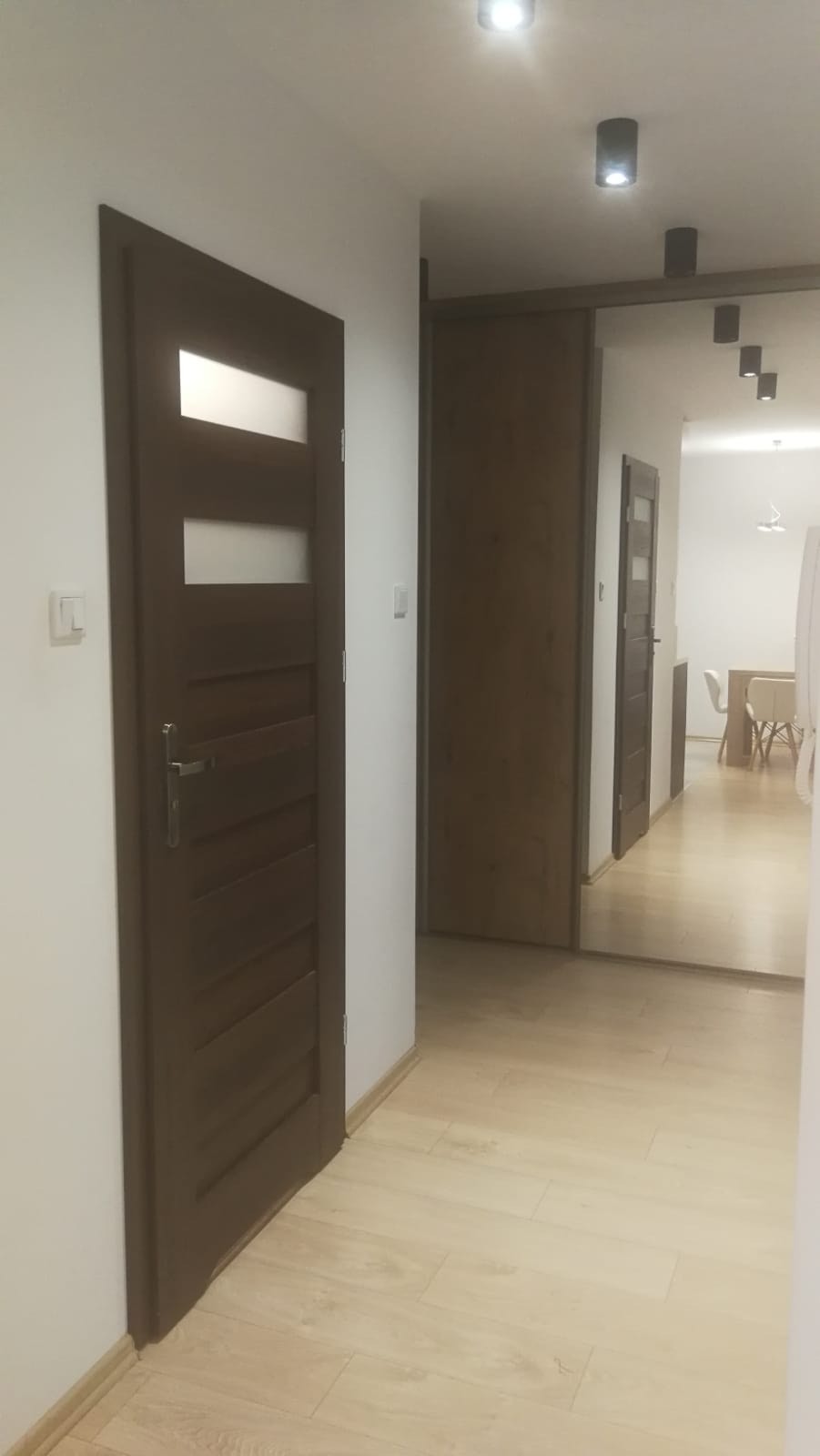 Wnętrze mieszkania z widokiem na drewniane drzwi z trzema przeszkleniami i podłogą z paneli w odcieniach beżu, oświetlone punktowo.