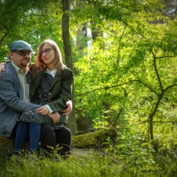 Para w leśnej scenerii: mężczyzna w kaszkiecie obejmuje kobietę w okularach siedzącą na pniu drzewa, otoczona zielenią.