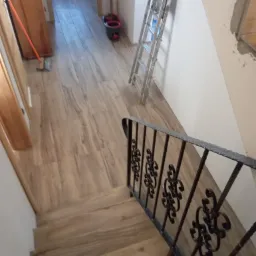 Nowe drewnopodobne panele podłogowe ułożone w korytarzu i na schodach, widok z g&oacute;ry na ozdobną, metalową barierkę schodową, drabina oparta o ścianę w tle.