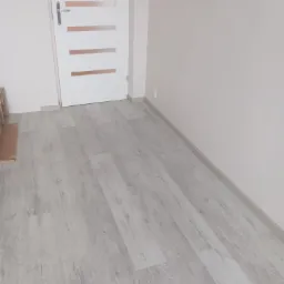 Jasnoszara podłoga panelowa w nowym wnętrzu, z białą, nowoczesną stolarką drzwiową i listwami przypodłogowymi. Świeżo wykończone, minimalistyczne pomieszczenie.