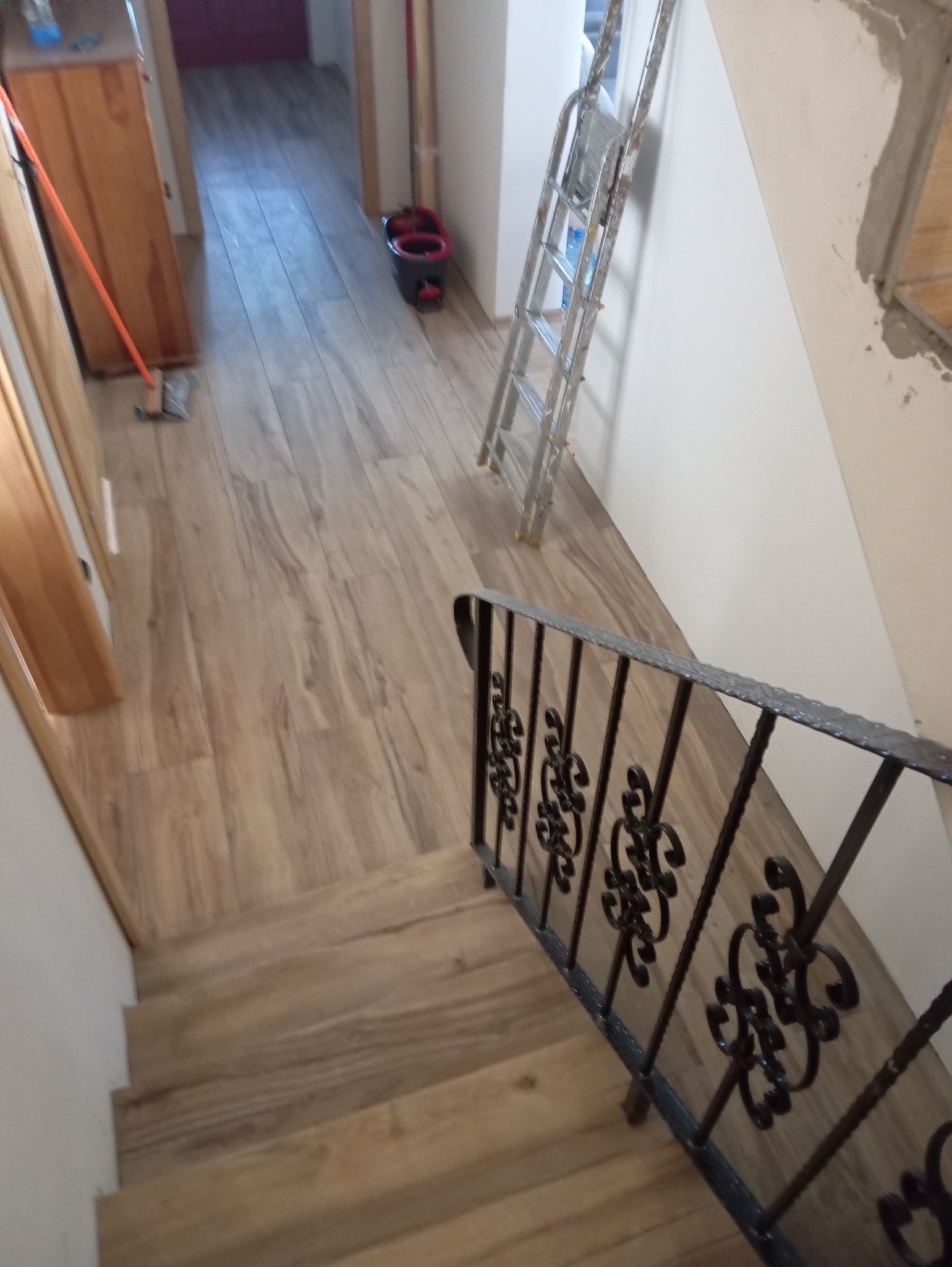 Nowe drewnopodobne panele podłogowe ułożone w korytarzu i na schodach, widok z góry na ozdobną, metalową barierkę schodową, drabina oparta o ścianę w tle.