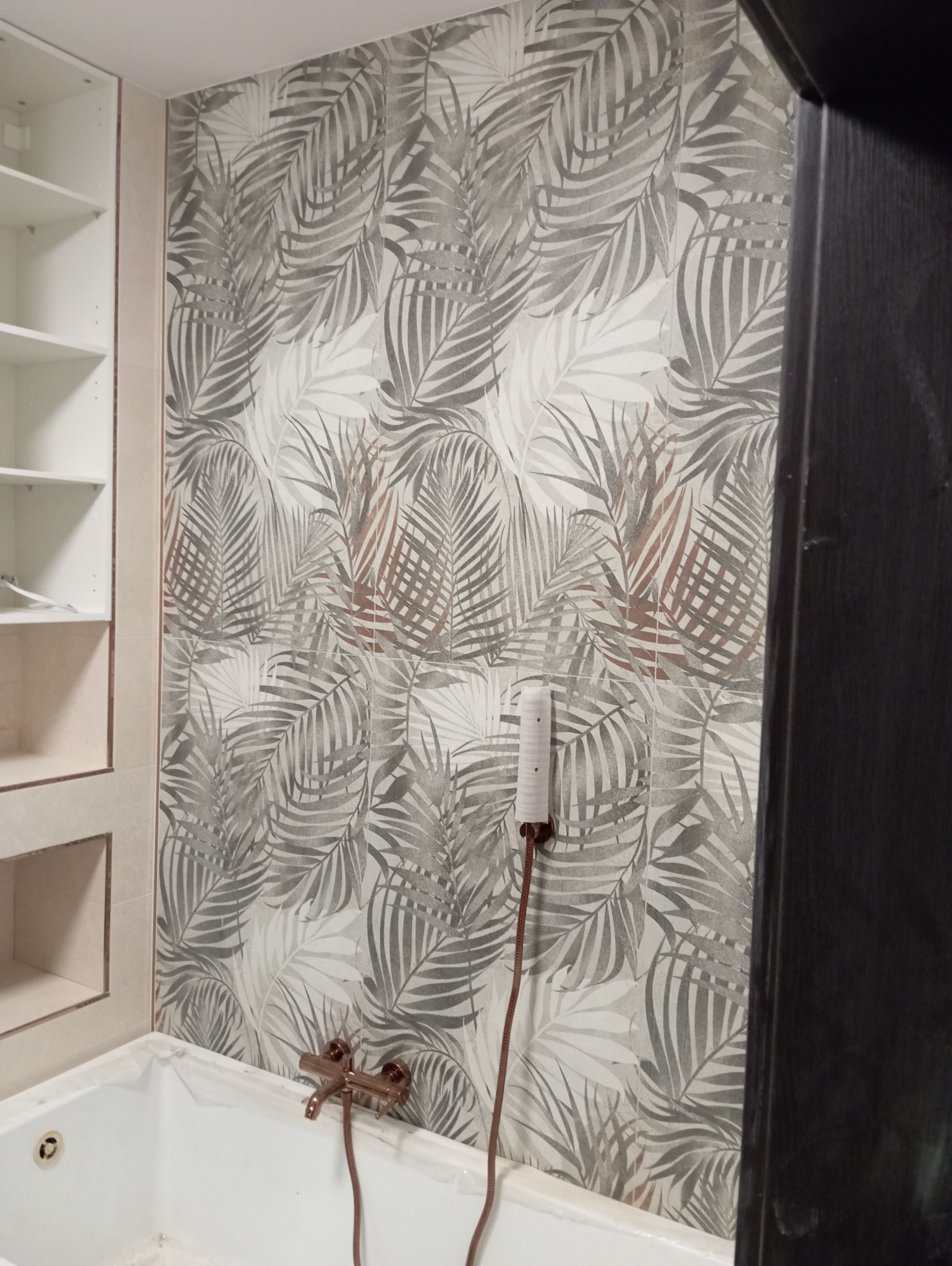 Wykończona łazienka z wanną, baterią w kolorze różowego złota i ścianami wyłożonymi płytkami z motywem liści palmowych w odcieniach szarości.