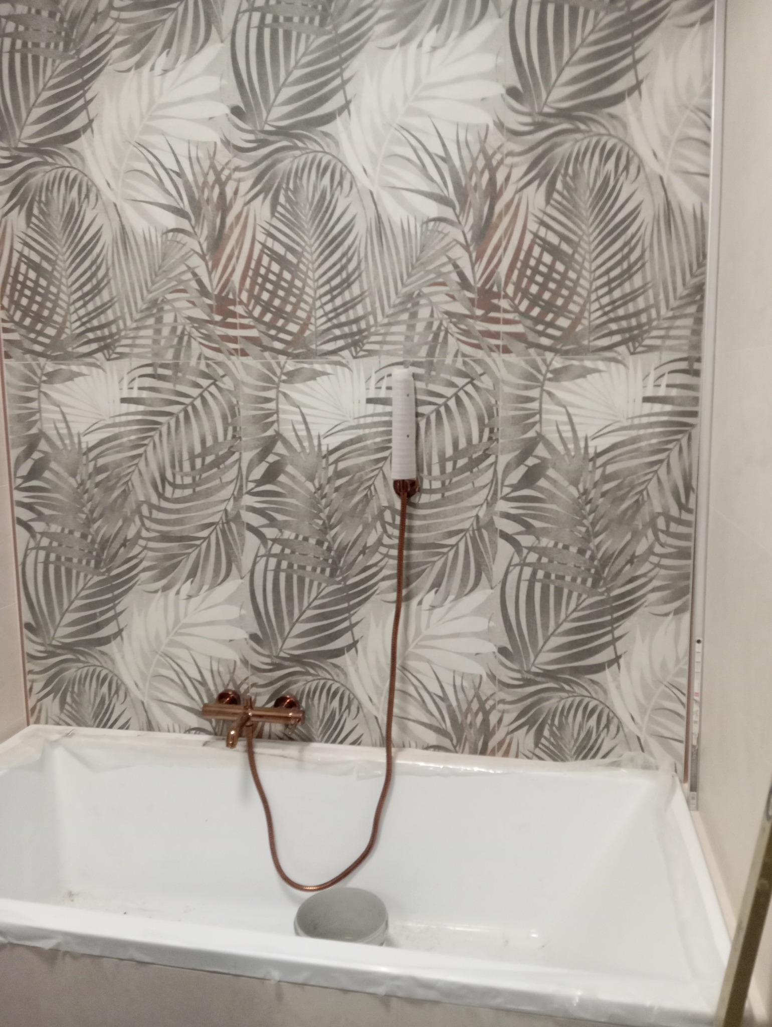 Biała wanna z baterią w kolorze miedzi i słuchawką prysznicową na tle ściany wyłożonej płytkami z motywem liści palmowych w odcieniach szarości.