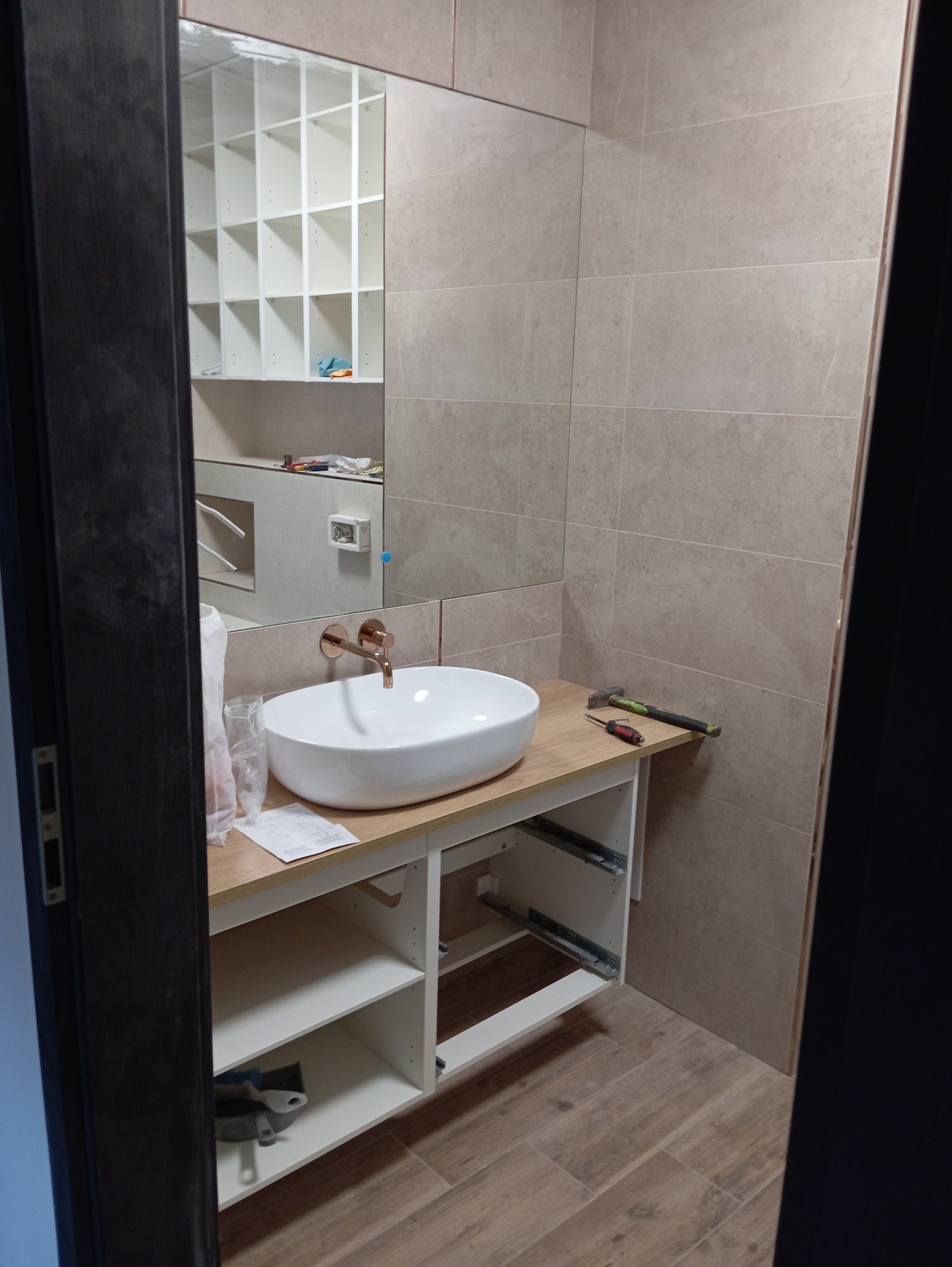 Montaż umywalki nablatowej z baterią w kolorze różowego złota na drewnianym blacie w łazience z beżowymi płytkami; widoczne narzędzia, szafka pod umywalką w trakcie montażu.