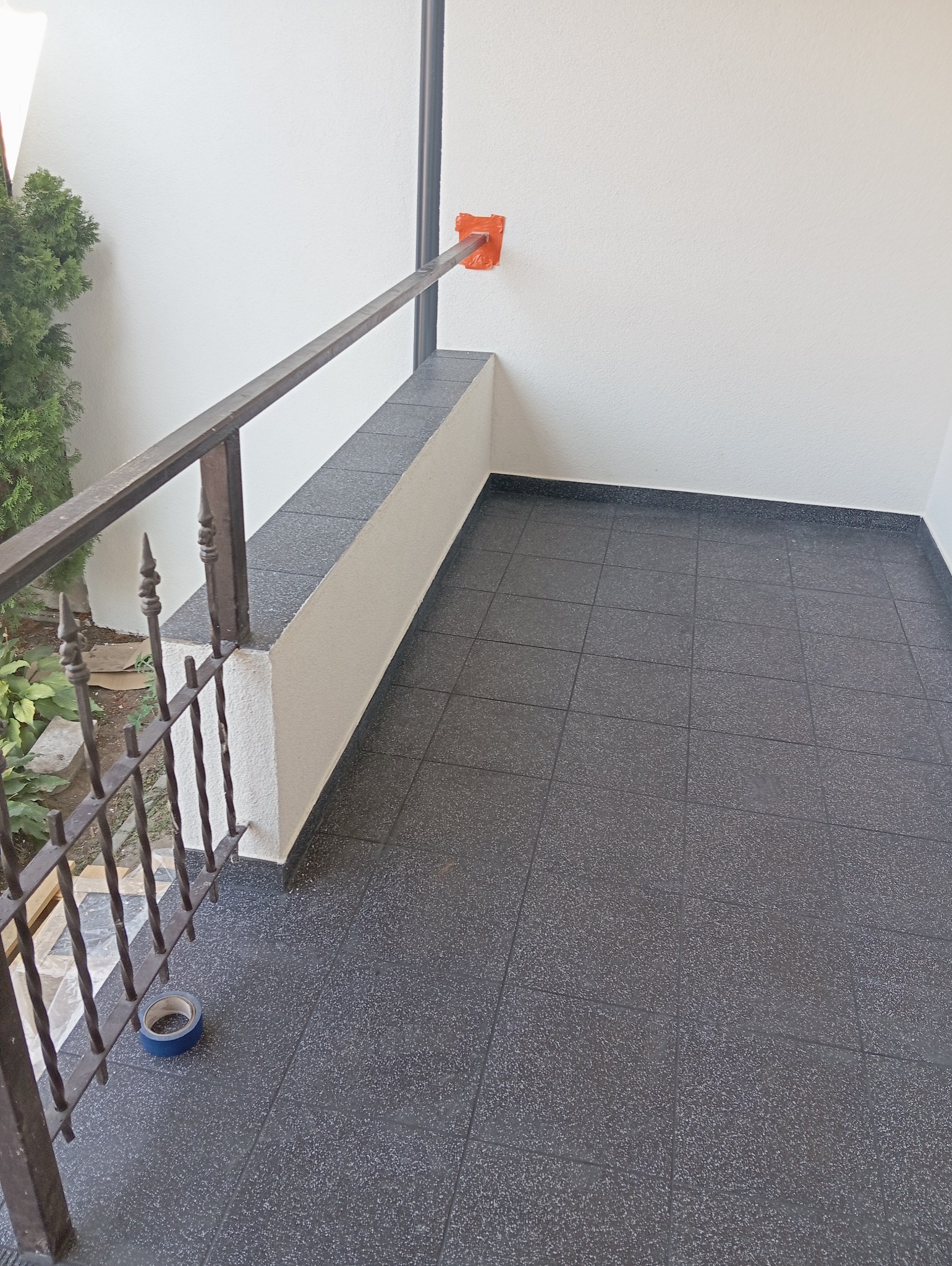 Wykończony balkon z ciemnoszarą, kwadratową podłogą z płytek i metalową balustradą. Pomarańczowa taśma zabezpieczająca wokół elementu mocującego balustradę do ściany. Na podłodze leży rolka...