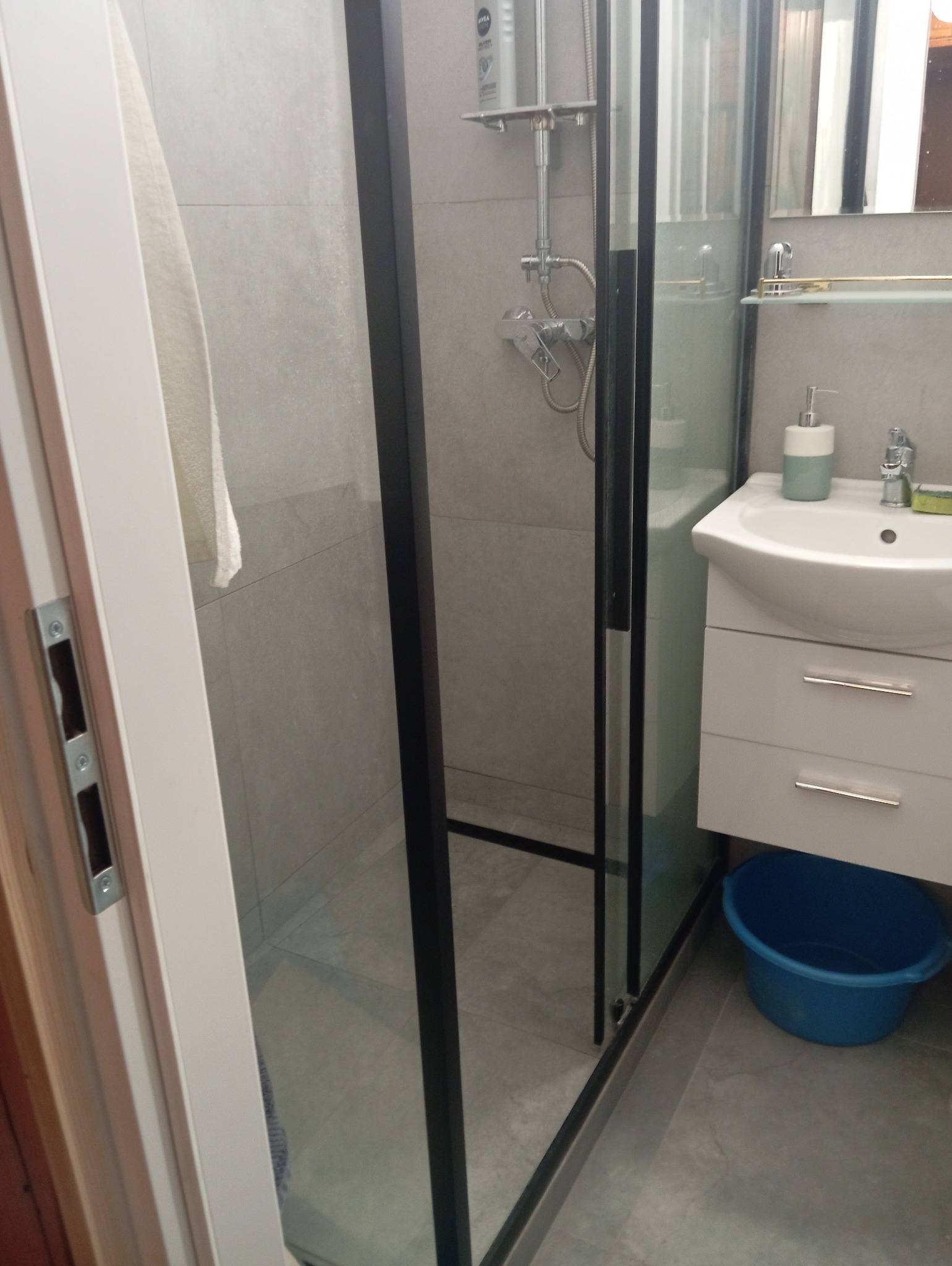 Nowoczesna kabina prysznicowa z czarnymi profilami, widoczna umywalka z szafką i niebieska miska na podłodze, szare płytki na ścianach i podłodze.