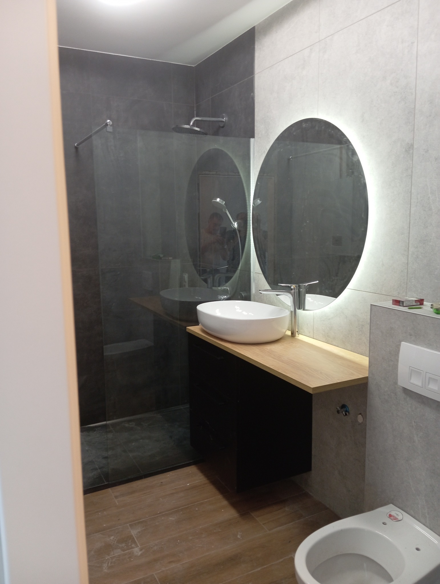 Nowoczesna łazienka z kabiną prysznicową typu walk-in, umywalką nablatową na drewnianym blacie, podświetlanym lustrem i bidetem. Stonowana kolorystyka szarości i brązu.