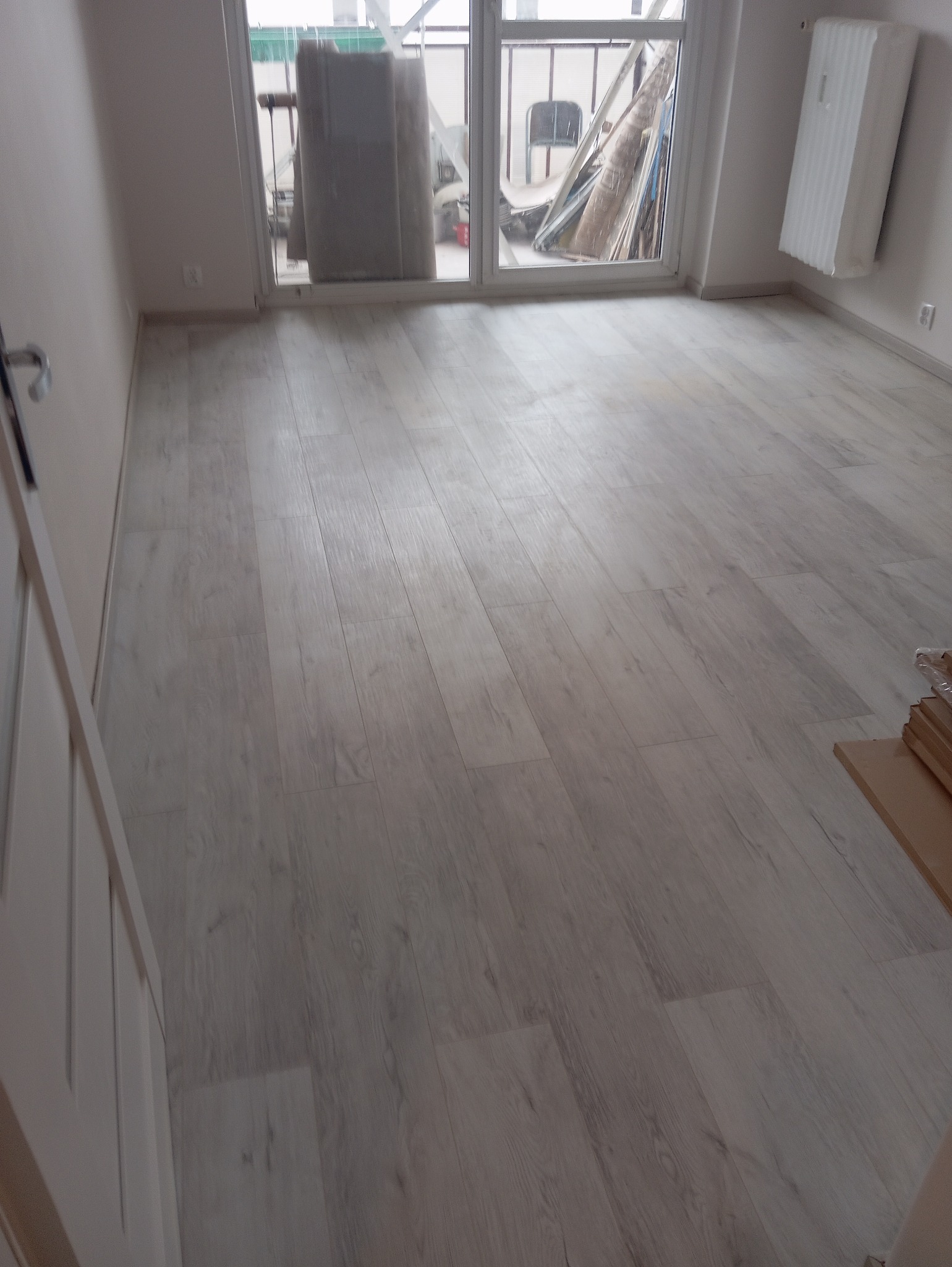 Jasna, drewnopodobna podłoga z panelami ułożonymi wzdłuż pokoju, widok na okno balkonowe i grzejnik na ścianie. Częściowo widoczne drzwi.