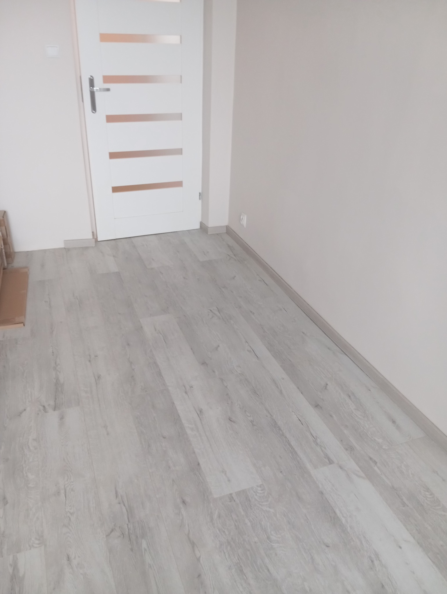 Jasnoszara podłoga panelowa w nowym wnętrzu, z białą, nowoczesną stolarką drzwiową i listwami przypodłogowymi. Świeżo wykończone, minimalistyczne pomieszczenie.