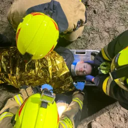 Ratownicy medyczni w kaskach stabilizują poszkodowaną kobietę z kołnierzem ortopedycznym na noszach, przykrytą złotą folią termiczną, na zewnątrz.