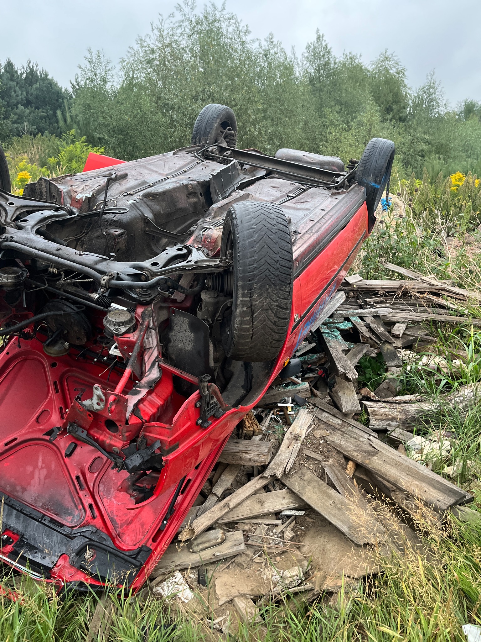 Przewrócony czerwony samochód osobowy na dachu, uszkodzony po wypadku, widoczne elementy zawieszenia, koła i karoseria wśród gruzu i trawy.