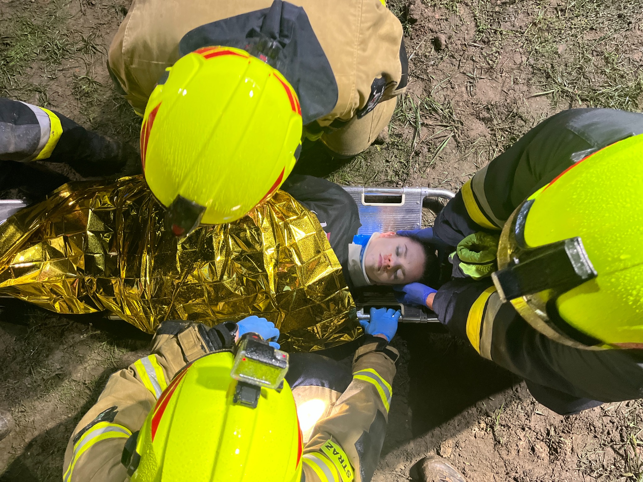 Ratownicy medyczni w kaskach stabilizują poszkodowaną kobietę z kołnierzem ortopedycznym na noszach, przykrytą złotą folią termiczną, na zewnątrz.