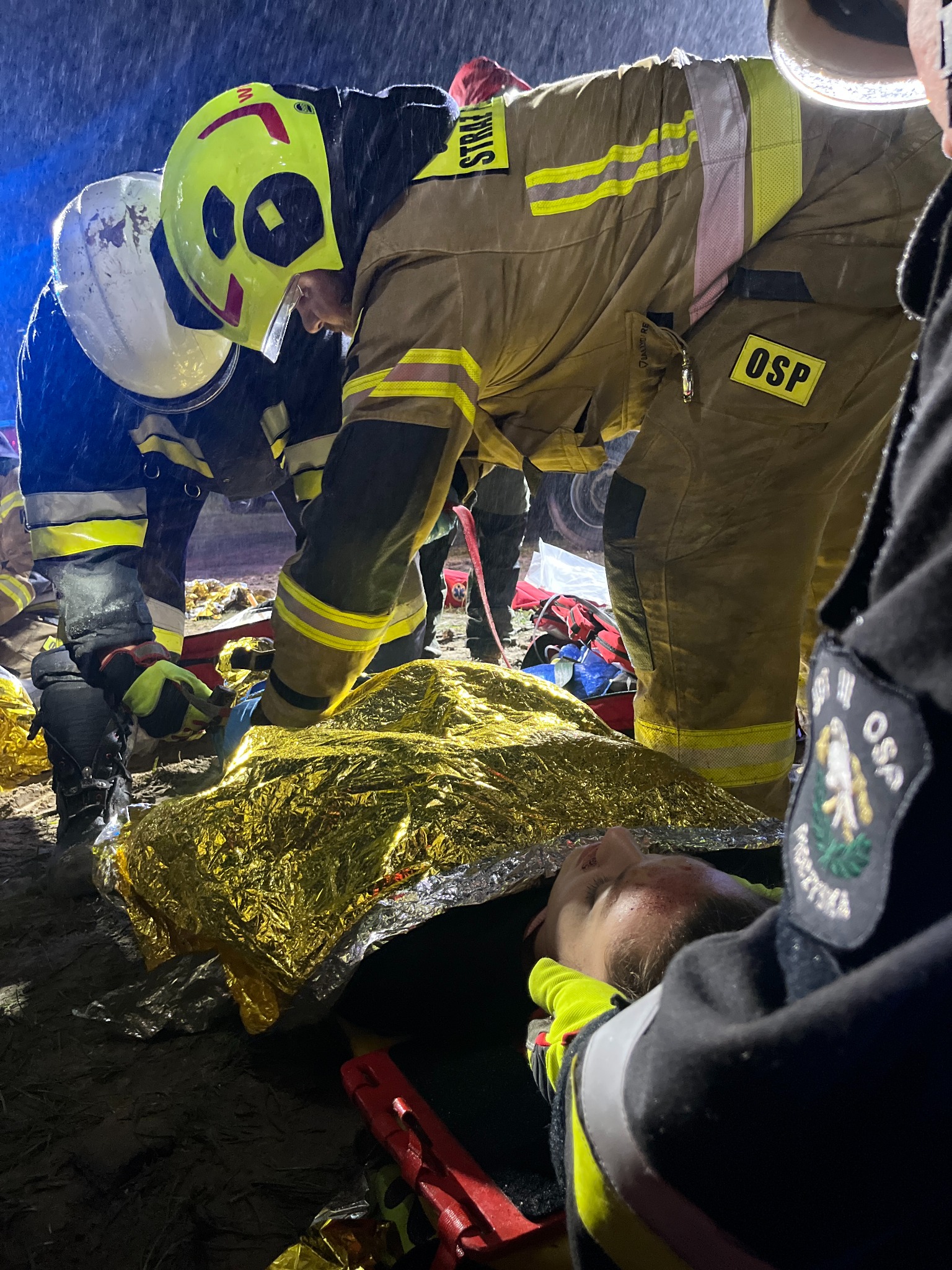 Ratownicy OSP udzielają pomocy poszkodowanemu, przykrytemu złotą folią termiczną, w warunkach nocnych podczas akcji ratunkowej.