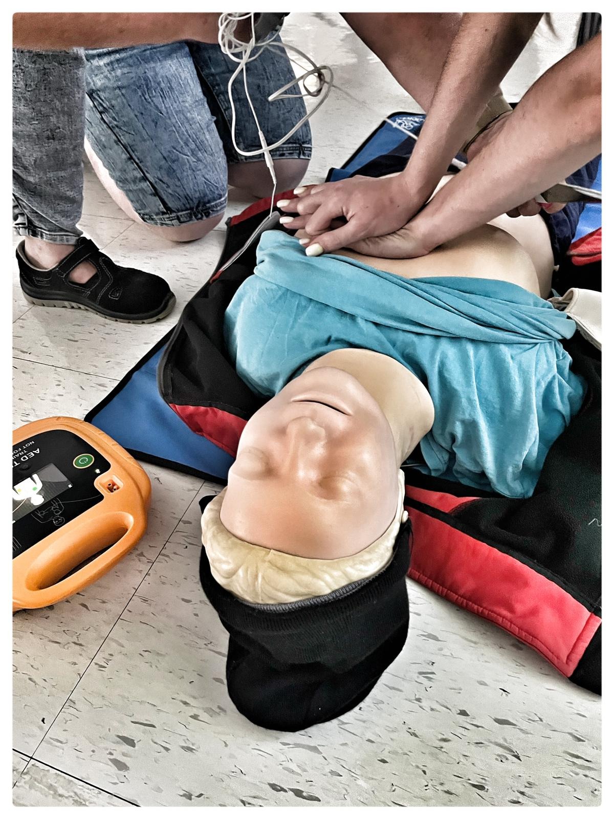 Instruktorzy wykonują resuscytację krążeniowo-oddechową na manekinie, obok leży defibrylator AED, widoczne stopy kursantów w tle.