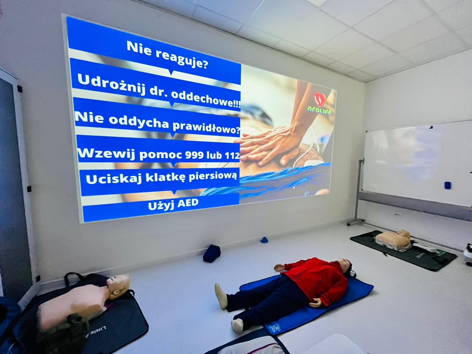 Sala szkoleniowa z manekinami do ćwiczeń resuscytacji krążeniowo-oddechowej; na ścianie wyświetlany jest slajd z instrukcją udzielania pierwszej pomocy.