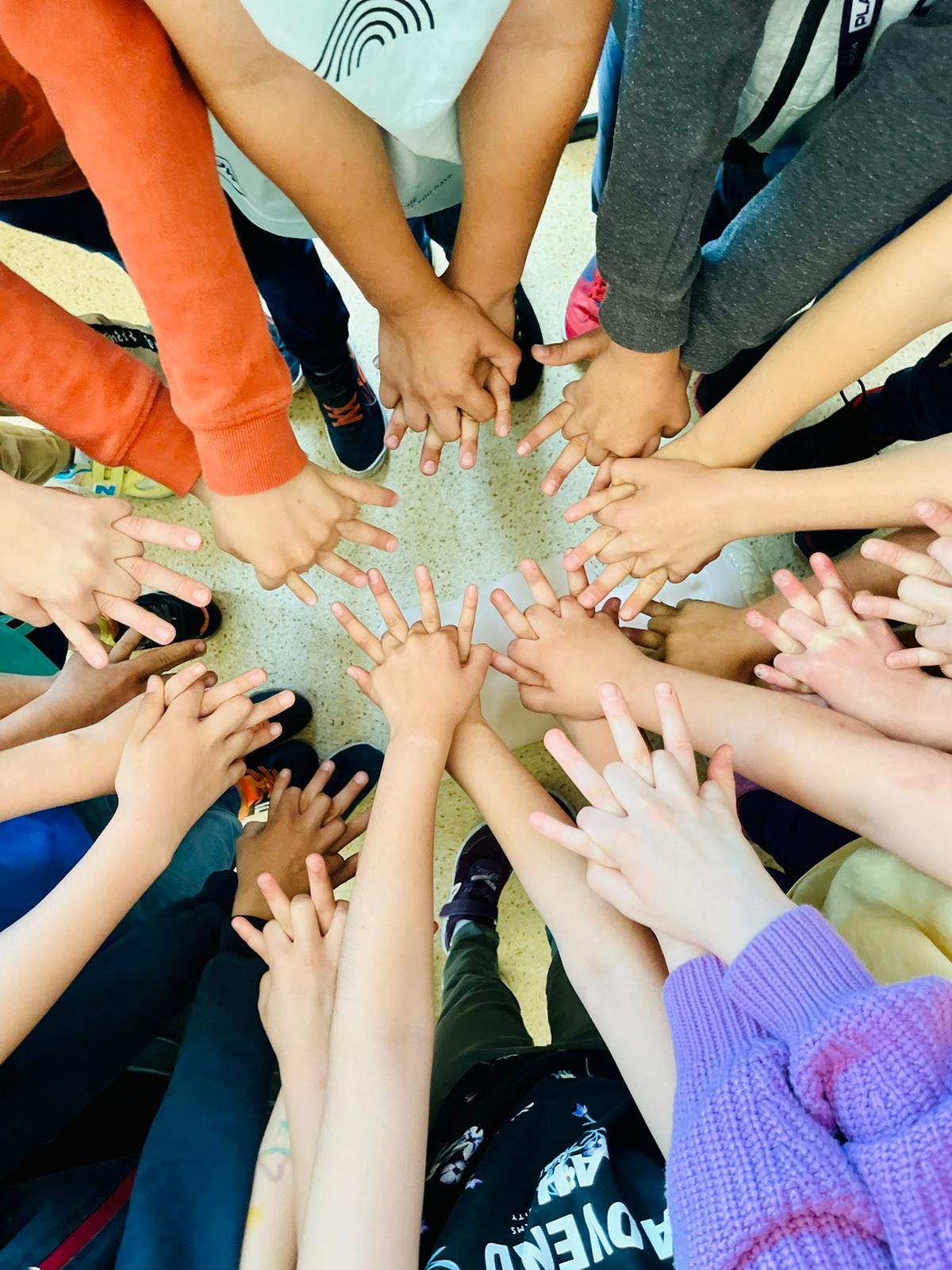 Grupka dzieci trzymających ręce w kręgu, unosząc dwa palce do góry, symbolizując zwycięstwo lub pokój, na tle podłogi w sali lekcyjnej.