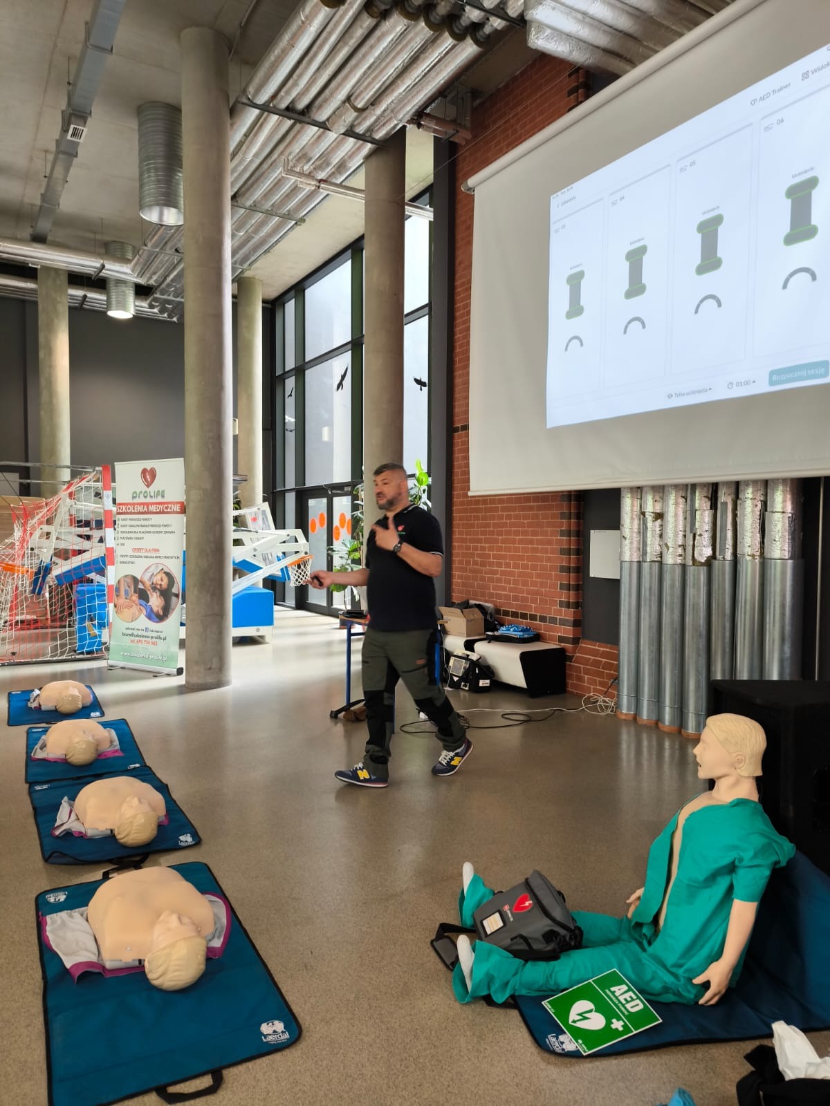 Instruktor prowadzi szkolenie z pierwszej pomocy, używając manekinów do resuscytacji i prezentacji multimedialnej z wizualizacją etapów resuscytacji krążeniowo-oddechowej; na podłodze leżą manekiny...