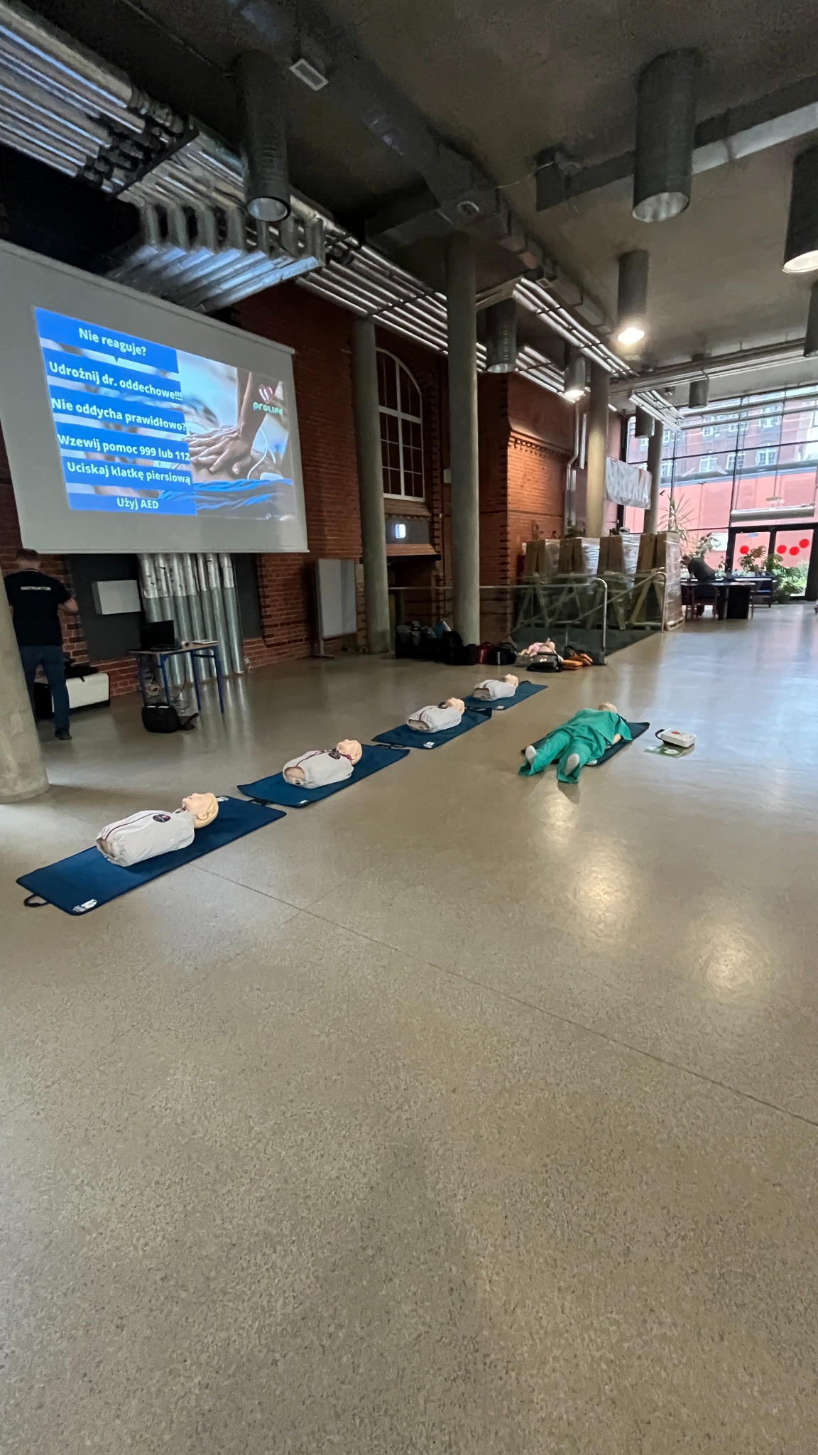 Sala szkoleniowa z manekinami do nauki pierwszej pomocy ułożonymi na matach; na ścianie wyświetlany slajd prezentacji z instrukcjami.