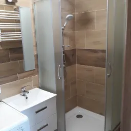 Nowoczesna łazienka z kabiną prysznicową narożną, umywalką z szafką w kolorze białym oraz pralką, wykończona płytkami imitującymi drewno z poziomym pasem w ciemniejszym odcieniu.