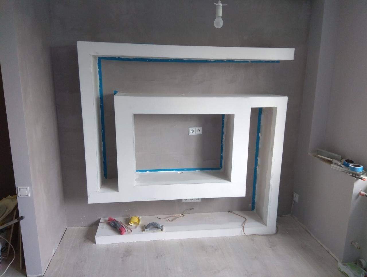 Nierówna ściana z surowym tynkiem, biała, geometryczna konstrukcja z płyty karton-gips zabezpieczona niebieską taśmą malarską, widoczne gniazdka elektryczne, narzędzia na podłodze.