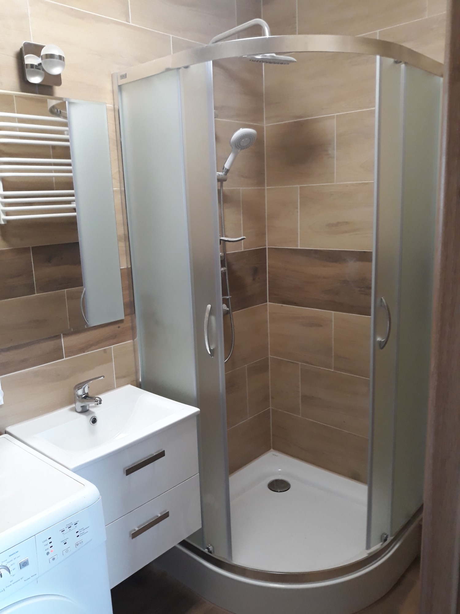 Nowoczesna łazienka z kabiną prysznicową narożną, umywalką z szafką w kolorze białym oraz pralką, wykończona płytkami imitującymi drewno z poziomym pasem w ciemniejszym odcieniu.