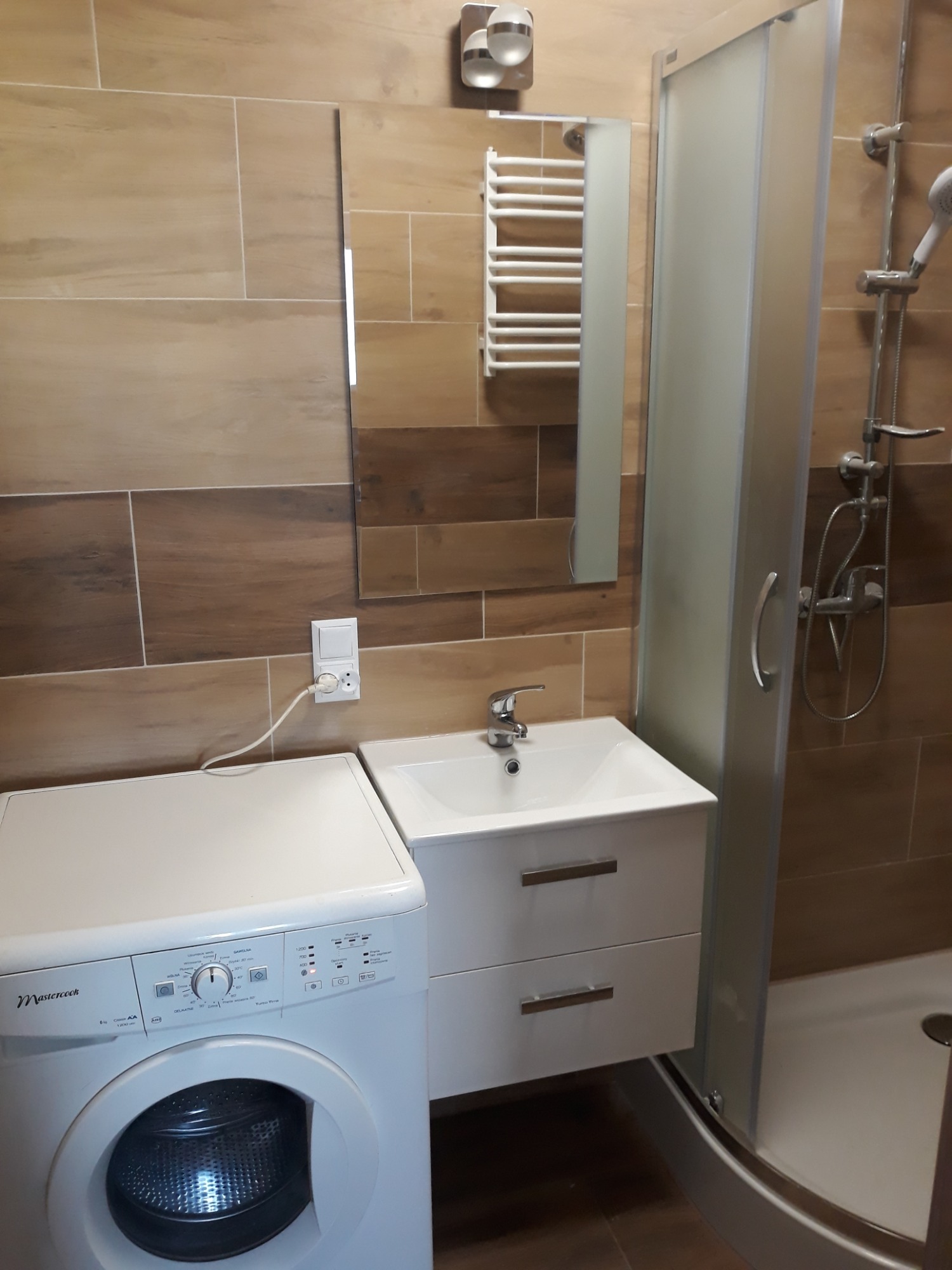 Ukończona łazienka z kabiną prysznicową, umywalką z szafką i pralką, wykończona płytkami imitującymi drewno, z grzejnikiem drabinkowym na ścianie.