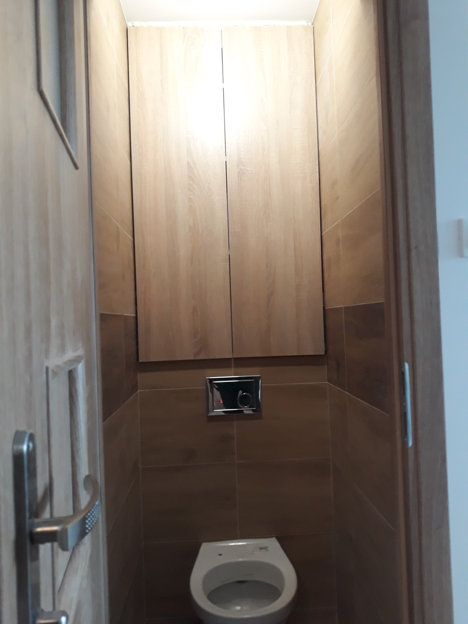 Mała łazienka z brązowymi płytkami, podwieszaną toaletą i szafką nad nią, widziana zza uchylonych drzwi.