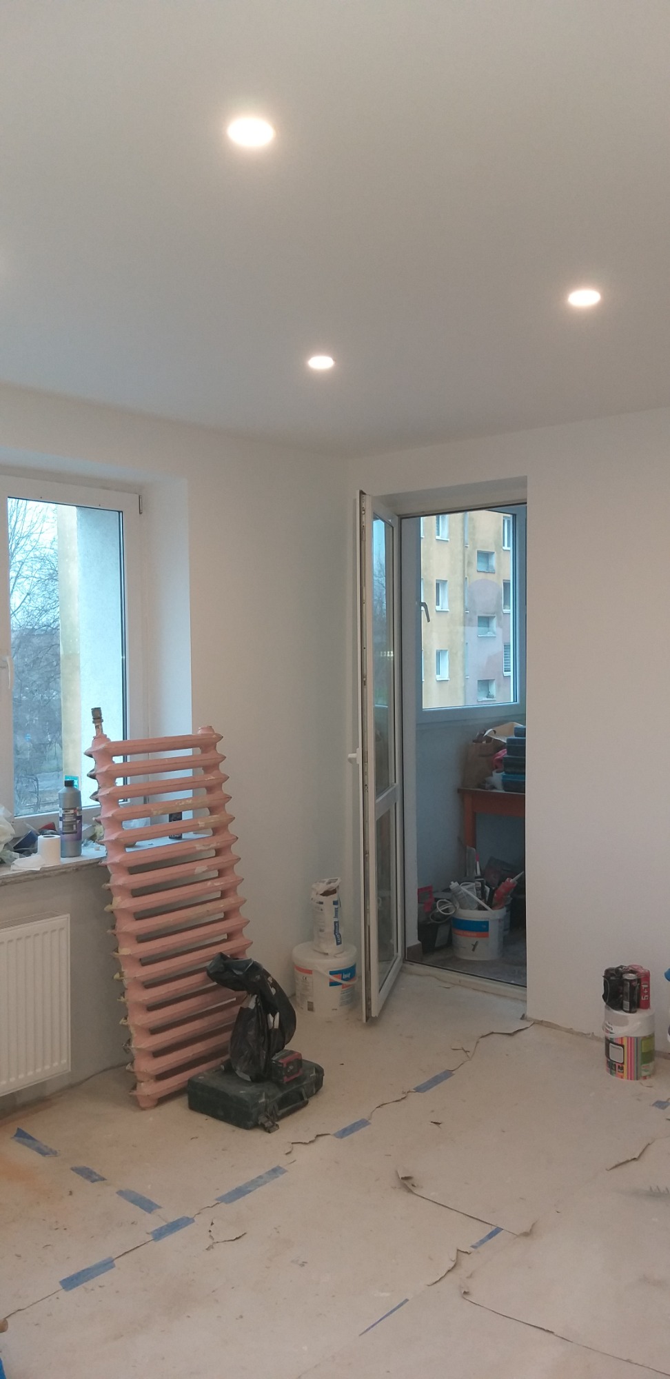 Pokój w trakcie remontu: świeżo pomalowane białe ściany, odnowiony różowy grzejnik oparty o ścianę, widoczne ślady taśmy malarskiej na podłodze, otwarte drzwi balkonowe z widokiem na budynek...